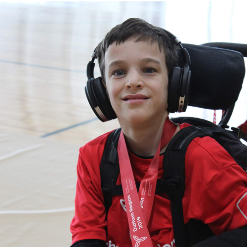 Young boy wearing headphones smiles from his wheelchair, with a medal around his neck.