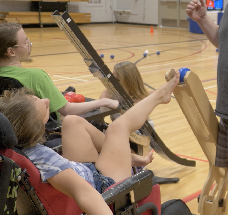 A young girl pushes a Boccia ball down a ramp with her right foot